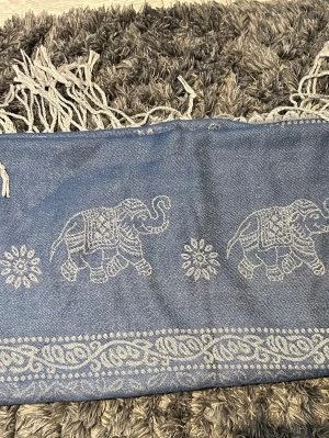 Blå sjal med elefantmönster - Snygg blå sjal med vita elefanter och blommor invävda i tyget. Sjalen har dekorativa fransar i ändarna och ett detaljerat kantmönster. Perfekt accessoar för att lyfta din outfit med en bohemisk touch.