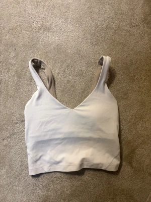 Beige tränings topp  från Lululemon - Säljer en vit träningstopp från Lululemon med breda axelband och v-ringad fram. Materialet är stretchigt och mjukt, perfekt för en bekväm känsla. Insidan är ljusare beige och plagget har en diskret logga på ryggen.