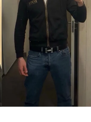 Svart hoodie från Hugo Boss med dragkedja - Snygg svart hoodie från Hugo Boss med gulddetaljer och dragkedja framtill. Hoodien har huva med snören och logga på bröstet. Insidan av huvan är beige. Perfekt för en avslappnad och trendig stil.