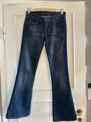 låga bootcut jeans med röda sömmar - Snygga mörkblå low waist bootcut jeans från rock&republic med röda kontrastsömmar och dekorativa detaljer på bakfickorna. Jeansen har klassisk femficksmodell och är tillverkade i denim. mjukt tyg och midjemåttet tvärs över är ca 37cm. Skriv för fler bilder