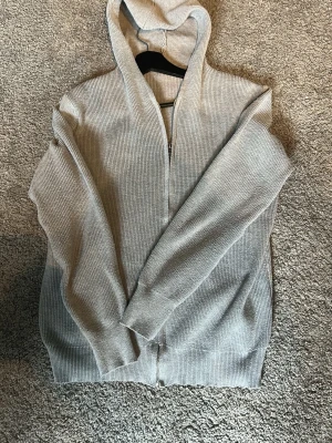  stickad hoodie med dragkedja - En stilren ljusgrå stickad hoodie med dragkedja framtill och huva. Tröjan har ribbad struktur och långa ärmar, perfekt för en avslappnad look. Passar bra till jeans.