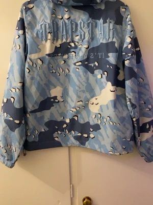 Blå camo vindjacka från Trapstar - Säljer en blå och vit camo vindjacka från Trapstar med huva och dragkedja framtill. Jackan har elastiska muddar vid ärmsluten och två fickor på framsidan. Trapstar-logga broderad på ryggen och patch på bröstet. Perfekt för streetwear-stilen.