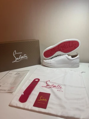 Vita Christian Louboutin sneakers - Säljer ett par vita sneakers från Christian Louboutin med ikonisk röd sula och coola nitar framtill. Skorna är i läder och har en klassisk låg siluett. Perfekta för dig som vill sticka ut med exklusiv stil och detaljer.
