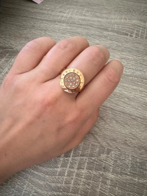 Rose färgad ring med stenar från LenMer - Snygg rose färgad ring med rund platta dekorerad med gnistrande stenar i mitten. Ringen har en bred design och text ingraverad runt plattan. Perfekt statement piece för dig som gillar att sticka ut. Ringen är 17 cm i omkrets!