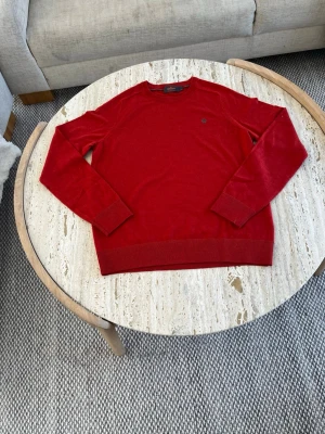 Röd stickad tröja med rund hals - Snygg röd stickad tröja med rund hals och långa ärmar. Tröjan har ribbade muddar vid ärmslut och nederkant samt en liten diskret logga på bröstet. Perfekt för dig som vill ha en enkel men stilren look.