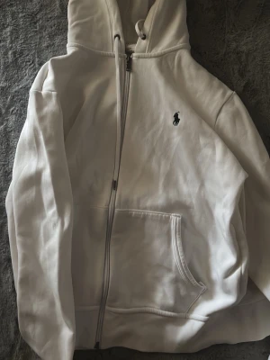 Vit hoodie från Polo Ralph Lauren - Säljer en vit hoodie från Polo Ralph Lauren med dragkedja, huva och känguruficka. Klassisk logga broderad på bröstet. Perfekt för dig som gillar stilrena plagg med premiumkänsla. Tillverkad i mjukt material för maximal komfort. Något år gammal, så kvalité inte på topp men funkar och är exakt som vanliga koftan. Pris kan diskuteras i DM.