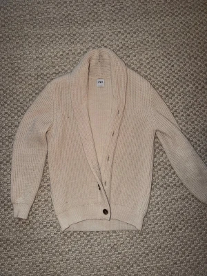 Beige stickad kofta från Zara - Mysig beige stickad kofta från Zara med grovstickad struktur och bred sjalkrage. Koftan har knappar framtill och ribbade muddar vid ärmslut och nederkant. Perfekt för lager på lager och en avslappnad look.