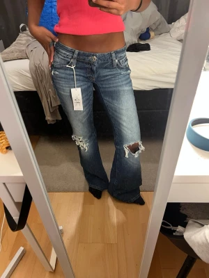 Blå slitna bootcut jeans med brodyr - Blåa lågmidjade bootcut jeans med slitningar och hål på knäna. Från Gina och aldrig använt 
