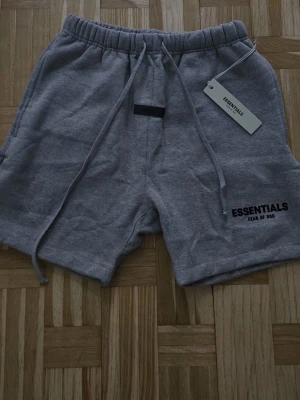 Grå shorts från Essentials Fear of God - Säljer ett par mörk grå shorts från Essentials Fear of God med elastisk midja och snörning.  Perfekta för chill dagar eller streetwear-stil. Mycket bra skick/ nytt skick. Använda endast 1 gång. Etiketterna finns kvar.