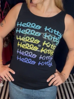 Hello Kitty svart linne - Snygg svart ärmlös topp med färgglada Hello Kitty-tryck i blått, gult och lila på framsidan. Toppen har en tight passform och är perfekt för dig som gillar statement-plagg med retrovibbar. Passar mig med storlek S/M.