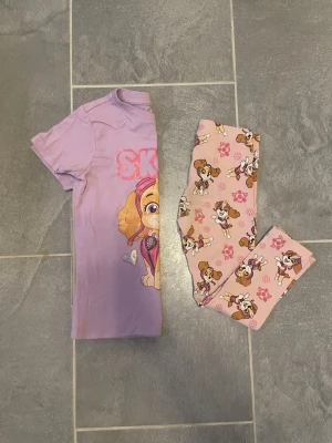 Rosa Paw Patrol pyjamasset för barn - Sött pyjamasset med kortärmad tröja i lila och matchande rosa leggings, båda med Skye från Paw Patrol. Tröjan har tryck framtill och leggingsen har ett all-over mönster med Skye och tassavtryck. Perfekt för dig som älskar Paw Patrol och vill ha något bekvämt att sova i.