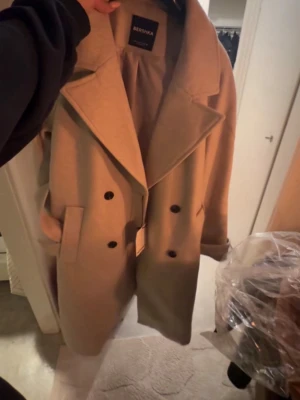 Beige dubbelknäppt kappa Bershka - Stilren beige kappa från Bershka med dubbelknäppning och breda slag. Kappan har två fickor framtill och en rak, något oversized passform. Perfekt för lager-på-lager och ger en trendig look till höst och vinter.