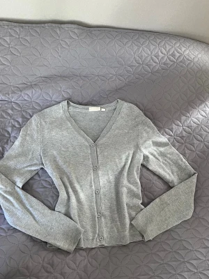 Grå kofta från KappAhl - Snygg och enkel grå kofta från KappAhl med v-ringning och knäppning framtill. Långärmad modell i mjukt material, perfekt att slänga över en t-shirt eller linne. Klassisk ribbad kant vid ärmslut och nederkant.