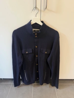 Mörkblå kofta från Fynch-Hatton, stl L - Snygg mörkblå kofta i superfine cotton från Fynch-Hatton. Modellen har ståkrage, bröstfickor med knappar, dragkedjor vid sidorna och träknappar längs hela framsidan. Perfekt för dig som gillar stilrena detaljer och praktiska fickor.