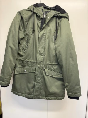 Olivgrön parkas med huva och foder - Snygg olivgrön parkas med dragkedja och tryckknappar framtill. Jackan har två stora fickor med lock, snedställda bröstfickor och justerbar huva med dragsko. Invändigt svart teddyfoder för extra värme. Perfekt för kalla dagar.