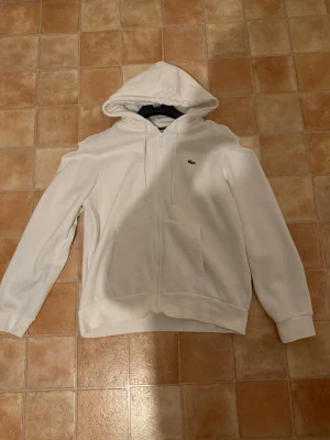 Vit hoodie med dragkedja från Lacoste - Snygg vit hoodie från Lacoste med dragkedja framtill och klassisk huva. Har två fickor på magen och den ikoniska gröna krokodilloggan på bröstet. Perfekt för en clean och stilren look. Materialet känns mjukt och bekvämt.