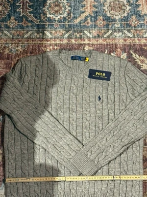 Grå kabelstickad tröja Polo Ralph Lauren - Kabelstickad grå tröja från Polo Ralph Lauren med rund halsringning och broderad logga på bröstet. Tröjan har långa ärmar och ribbade muddar vid ärmslut och nederkant. Perfekt för en stilren och klassisk look.