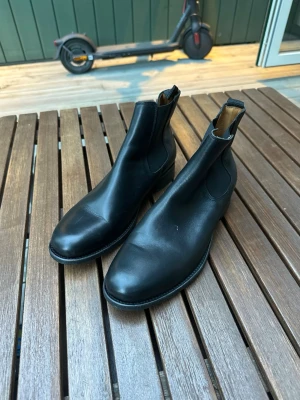 Svarta klassiska rizzo boots - Säljer ett par stilrena svarta Chelsea boots i slätt läder med elastiska paneler på sidorna och dragflik bak. Bootsens minimalistiska design gör dem enkla att matcha med olika outfits. Perfekta för dig som gillar tidlös och clean stil.