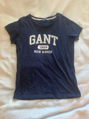 Marinblå GANT t-shirt med tryck - Snygg marinblå t-shirt från GANT med vit text 'GANT 1949 NEW HAVEN' på bröstet. Klassisk rund halsringning och korta ärmar. Perfekt för en avslappnad och stilren look. Den är i storlek xs på lappen, men skulle säga att den även passar som s