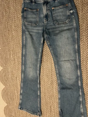 Blå bootcut jeans med fickor fram - Säljer ett par ljusblå bootcut jeans med raka ben och stora fickor fram. Jeansen har en klassisk tvättad look och är sydda i denim med kontrastsömmar. Perfekta för en avslappnad och trendig stil.