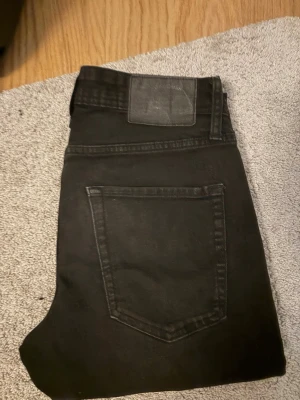 Svarta raka jeans med hög midja - Snygga svarta jeans med raka ben och hög midja. Klassisk femficksmodell med knappgylf och bälteshällor. Jeansen är i ett stretchigt denimtyg som sitter bekvämt och passar till många olika stilar.