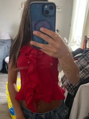Röd volang topp med omlottkänsla - Säljer en röd ärmlös topp med volangdetaljer och omlottkänsla. Tyget är strukturerat och stretchigt, vilket ger en snygg och unik look. Perfekt för dig som vill sticka ut lite extra! Toppen har en croppad passform och markerad midja.