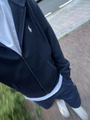 Marinblå hoodie från Polo Ralph Lauren - Snygg marinblå hoodie från Polo Ralph Lauren med dragkedja framtill, två fickor och klassisk vit logga på bröstet. Hoodien har huva med dragsko och ribbade muddar. Perfekt till chill vardagsstil och funkar året runt.