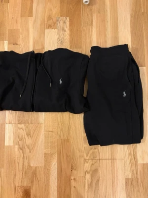 Svart hoodie och byxor från Polo Ralph Lauren - Säljer ett svart set med zip up hoodie och matchande byxor från Polo Ralph Lauren. Båda plaggen har den klassiska loggan broderad på bröstet och benet. Hoodien har dragkedja och snören i huvan, byxorna har resår i midjan och sidofickor. Koftan är i helt perfekt skick utan defekter. Byxorna är densamma förutom sista bilden som visar en liten defekt på snörena vid byxorna. Koftan är storlek M och Byxorna L. Pris kan diskuteras