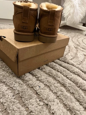 Bruna UGG fårskinnsstövlar - Säljer ett par klassiska bruna UGG boots i mocka med mjukt fårskinnsfoder. De har en rund tå och en stadig sula, perfekta för kalla dagar. UGG-loggan syns tydligt bak på hälen. Kommer med originalkartong.