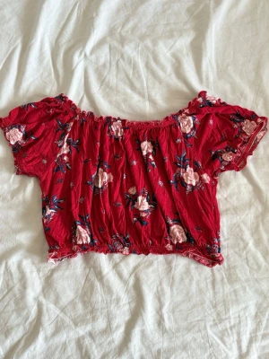Röd blommig croppad offshoulder topp - Söt röd croppad topp med blommigt mönster och offshoulder-design. Toppen har korta puffärmar och resår vid axlar och midja.
