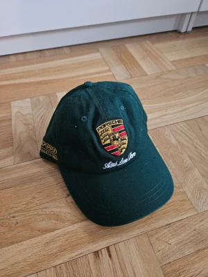 Mörkgrön Porsche x Aime Leon Dore keps - Snygg mörkgrön keps från Aime Leon Dore x Porsche collaben med broderad Porsche-logga framtill. Kepsen har böjd skärm och detaljer i gult och rött på sidan och i fram. Tillverkad i bomullstyg och justerbar baktill. Mörkgrön.