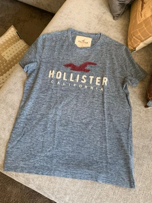 Blåmelerad Hollister t-shirt - Snygg blåmelerad t-shirt från Hollister med klassisk logga och texten 'Hollister California' framtill. T-shirten har rund hals och korta ärmar, perfekt för en avslappnad och stilren look.