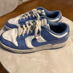 Nike Dunk Low blå och vita sneakers - Nike Dunk Low sneakers i blått och vitt med klassisk swoosh på sidan. Skorna har vita snören, perforerad tåbox och vadderad plös. Ovandelen är i läder och insidan är mjukt fodrad för extra komfort. Perfekt för dig som gillar streetwear och sportig stil pris och frågor går att diskuteras via dm använda max 5 gånger 