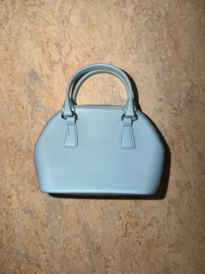 Ljusblå minihandväska med dragkedja - Söt ljusblå minihandväska i syntetmaterial med rundad form och två korta handtag. Väskan har dragkedja upptill och minimalistisk design, perfekt för att bära småsaker. Enkel och stilren look som passar till många outfits.