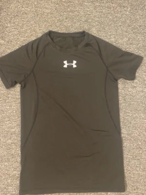 Svart träningströja Under Armour - Säljer en svart, kortärmad träningströja från Under Armour. Tröjan har en tight passform och är tillverkad i ett snabbtorkande funktionsmaterial. Loggan sitter framtill på bröstet i vitt. Perfekt för gymmet eller löpning.