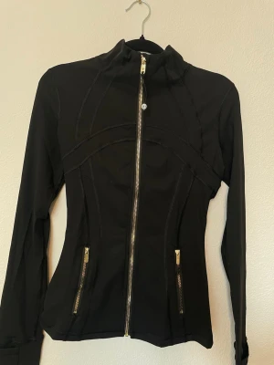Lululemon define  - Snygg svart ziptröja med figurnära passform och guldiga dragkedjor framtill och på fickorna. Hög krage och långa ärmar. Perfekt för dig som gillar sportig och stilren look. Materialet känns stretchigt och bekvämt.