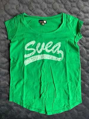 Grön t-shirt från Svea - Grön t-shirt från Svea med vitt tryck på bröstet. Klassisk rund halsringning och korta ärmar. 