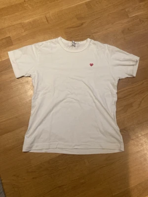 Vit t-shirt Comme des Garçons Play - Säljer en klassisk vit t-shirt från Comme des Garçons Play med det ikoniska lilla röda hjärtat broderat på bröstet. T-shirten har rund hals och korta ärmar, tillverkad i mjuk bomull för en skön känsla. Perfekt till en clean och stilren look. Är en medium men sitter som en small.