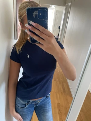Mörkblå t-shirt med logga - Snygg mörkblå t-shirt med korta ärmar och liten vit logga på bröstet. Klassisk passform och rund halsringning. Perfekt till jeans eller shorts för en enkel och stilren look.