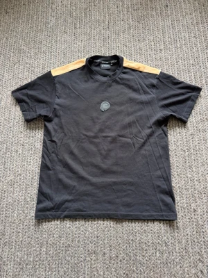 Svart t-shirt - Svart t-shirt från Vichy med beige axelpartier och rund hals. T-shirten har ett diskret märke på bröstet och är tillverkad i mjuk bomull. Perfekt för en avslappnad och stilren look.