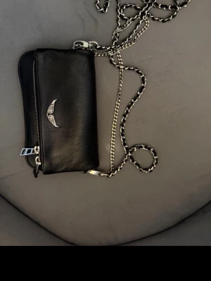 Svart handväska Zadig & Voltaire - Snygg svart handväska i äkta läder från Zadig & Voltaire. Väskan har en silverfärgad kedjerem, dekorativt vingemblem framtill och dragkedja med metall-detaljer. Perfekt för dig som gillar stilrena accessoarer. Väskan har endast en skavank som sitter på dragkedjan och det är en läderbit som åkt av.