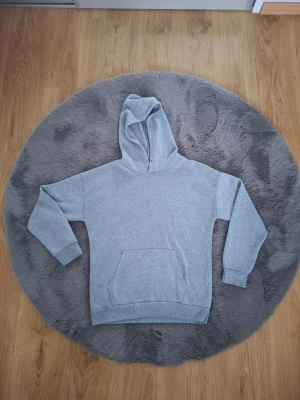 Ljusgrå basic hoodie med huva - En stilren ljusgrå hoodie med klassisk huva och stor magficka framtill. Hoodien har ribbade muddar vid ärmar och nederkant, samt en avslappnad passform. Perfekt för chill dagar eller när du vill ha en enkel och clean look.