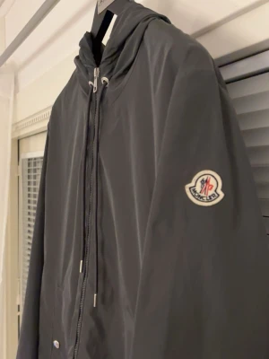 Svart vindjacka från Moncler storlek 3 - Snygg svart vindjacka från Moncler med huva och dragsko. Jackan har dragkedja framtill, två fickor med tryckknappar och Moncler-logga på vänster ärm. Tillverkad i ett lätt och smidigt material som skyddar mot vind.