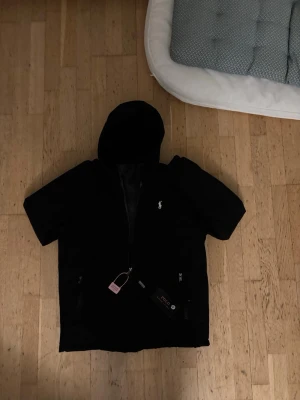 Polo Ralph lauren vindjacka - Snygg svart vindjacka med huva och dragkedja framtill. Jackan har två fickor med dragkedja och en liten vit logga på bröstet. Materialet ser ut att vara vattenavvisande och jackan har en sportig look. Perfekt för blåsiga dagar. Storlek XL i barn storlek, sitter som M i vuxen storlek. Pris ej hugget i sten
