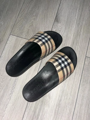 Burberry slides med rutigt band - Säljer ett par snygga slides från Burberry med klassiskt beige, svart och vitt rutigt band över foten. Sulan är svart och har en diskret Burberry-logga på sidan. Tillverkade i slitstark syntet, perfekta för en trendig och avslappnad stil.
