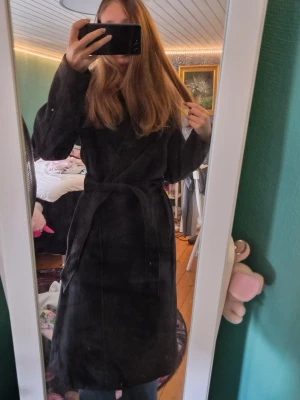 Svart lång kappa från H&M - Stilren svart lång kappa från H&M med knytskärp i midjan och klassisk krage. Kappan har en rak passform och är helfodrad för extra komfort. Perfekt för dig som vill ha en enkel men snygg look till hösten eller vintern.