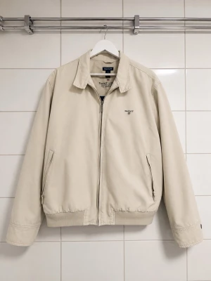 GANT JACKA  - Stilren beige Jacka  från GANT med dragkedja framtill, två snedställda fickor och broderad logga på bröstet. Modellen är 186 cm. 