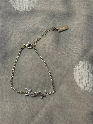 Silverfärgat YSL armband - Stilrent armband i silverfärg med YSL-logga framtill och tunn kedja. Justerbar längd med liten bricka där det står Saint Laurent Paris. Perfekt accessoar för att lyfta din outfit.