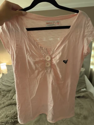 Ljusrosa topp från Roxy - Söt ljusrosa topp från Roxy med korta ärmar och v-ringning. Toppen har tre dekorativa knappar framtill och en liten broderad logga på bröstet. Perfekt för en avslappnad och trendig look.