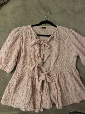 Ljusrosa blus med knytningar - Söt ljusrosa blus i krinklat tyg med tre knytningar framtill. Blusen har trekvartsärm och en lätt utsvängd passform nedtill. Perfekt för en trendig och avslappnad look.
